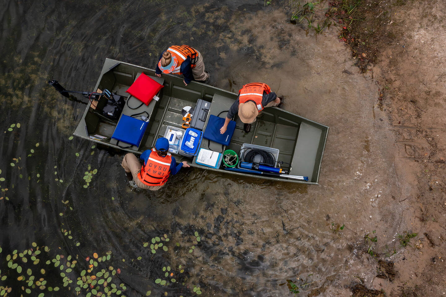 Three individuals in safety vests are working in a small boat on a waterway with floating vegetation, surrounded by muddy banks.